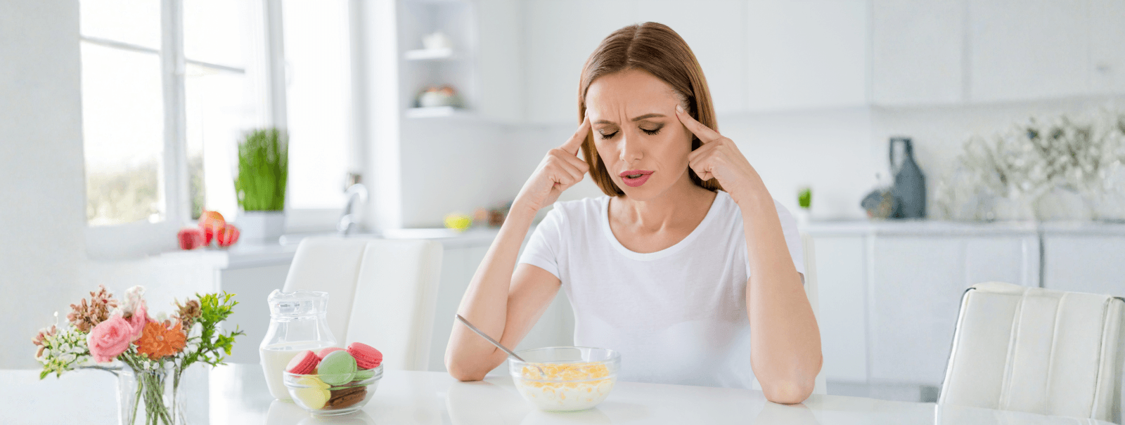 Woman sitting at a kitchen table holding her temples, appearing fatigued or stressed, with a bowl of cereal and sugary snacks nearby in a bright, modern kitchen